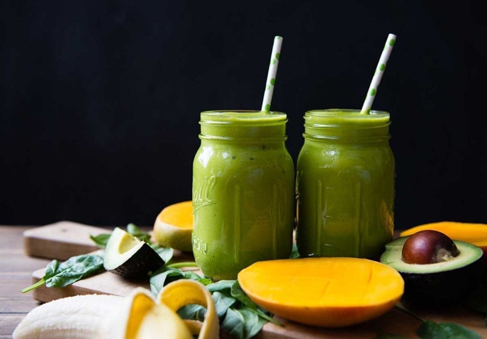 Two mason jars full of green smoothie surrounded by fresh avocado and bananas.