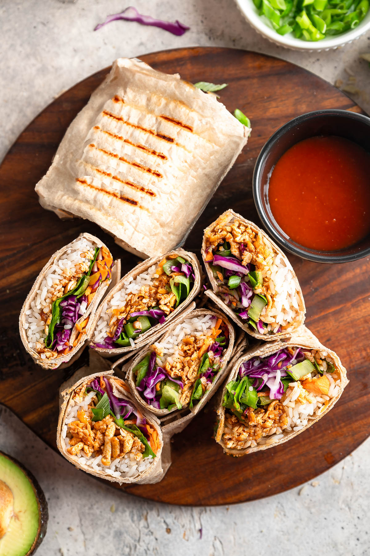 A wooden board displays a vibrant arrangement of buffalo shredded tofu wraps with buffalo sauce on the side. 