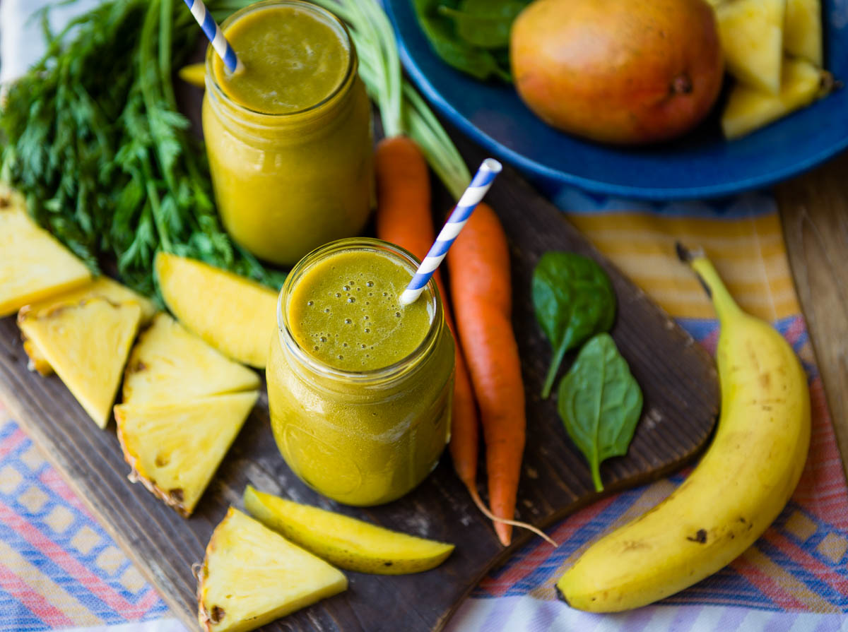 Carrot greens smoothie with papers straws, surrounded by fresh carrots, mango and pineapple slices.