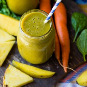 Carrot greens smoothie in a mason jar with a paper straw next to carrot tops on table.