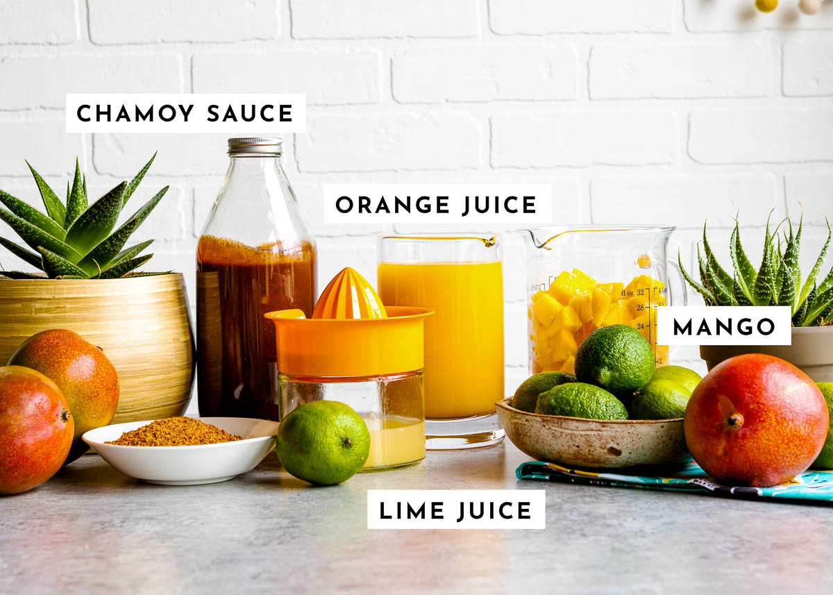 Labeled mangonada ingredients on countertop including chamoy sauce, orange juice, mango and limes.