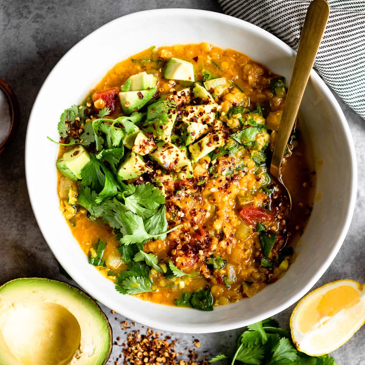 red lentil soup in a white bowl, topped with fresh avocado and a gold spoon.