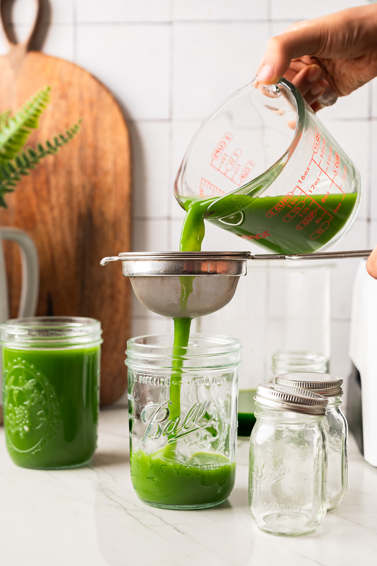 Green juice being poured through a fine mesh sieve into a glass jar