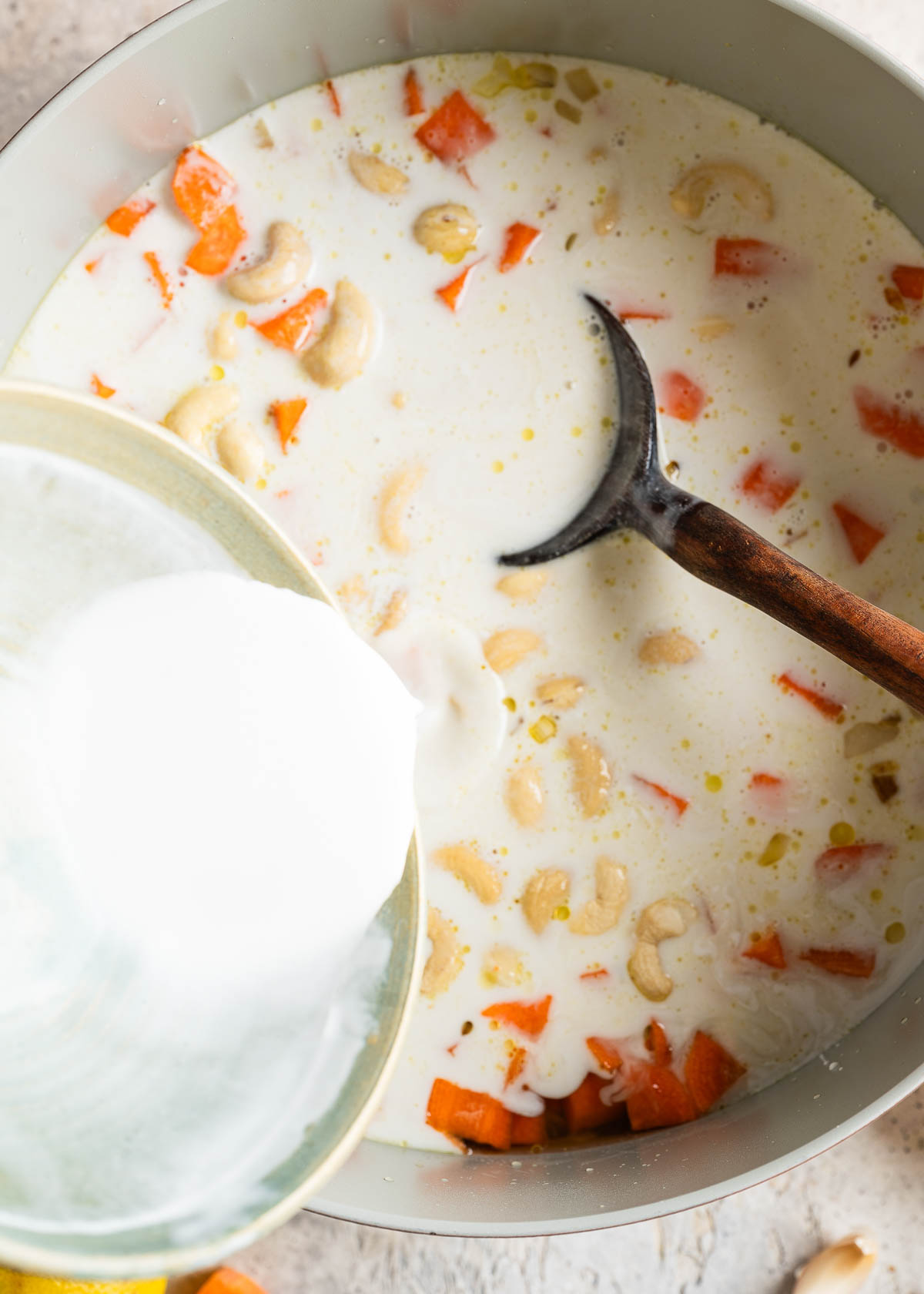Pouring coconut milk into pot with diced carrots, cashews and ginger.