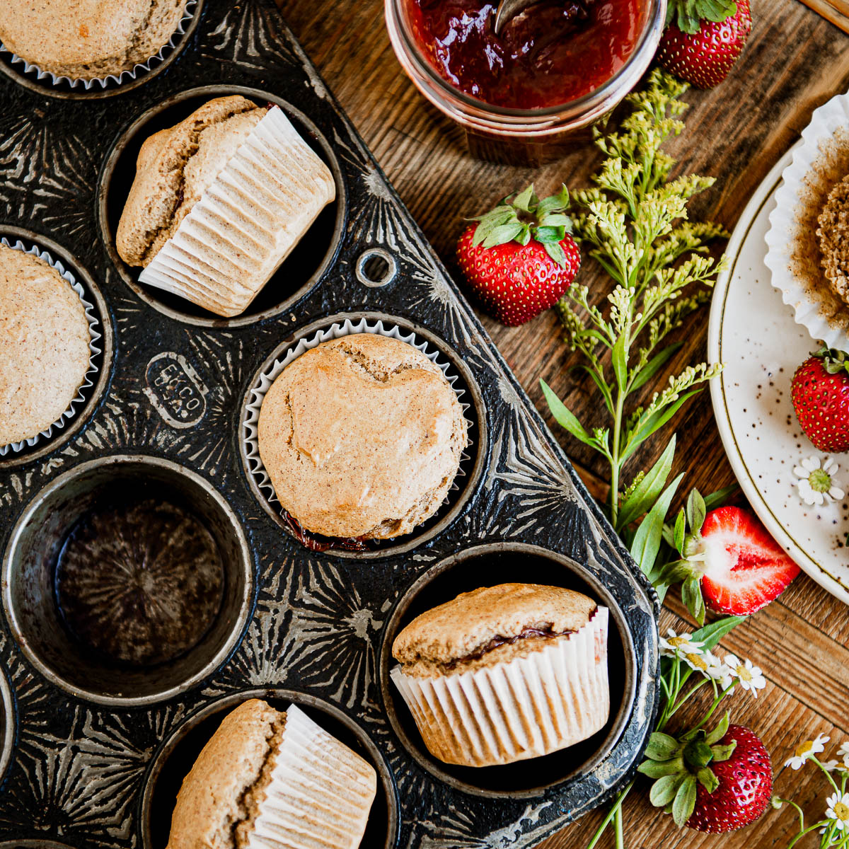 Antique black muffin tin with baked vegan muffins in white wrappers, next to strawberry jam.