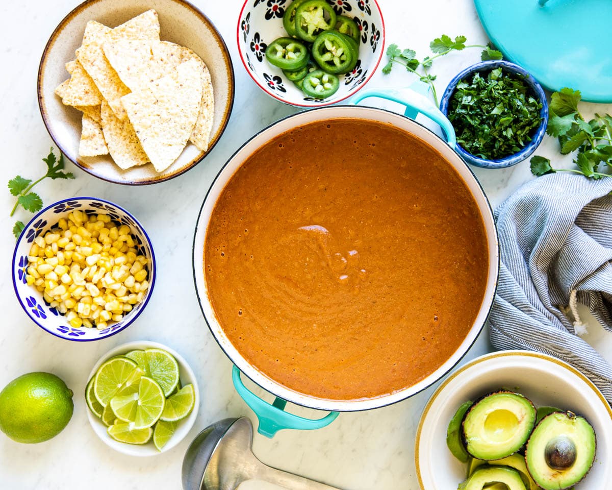 Healthy vegetarian tortilla soup in aqua stock pot with bowls of toppings surrounding it.